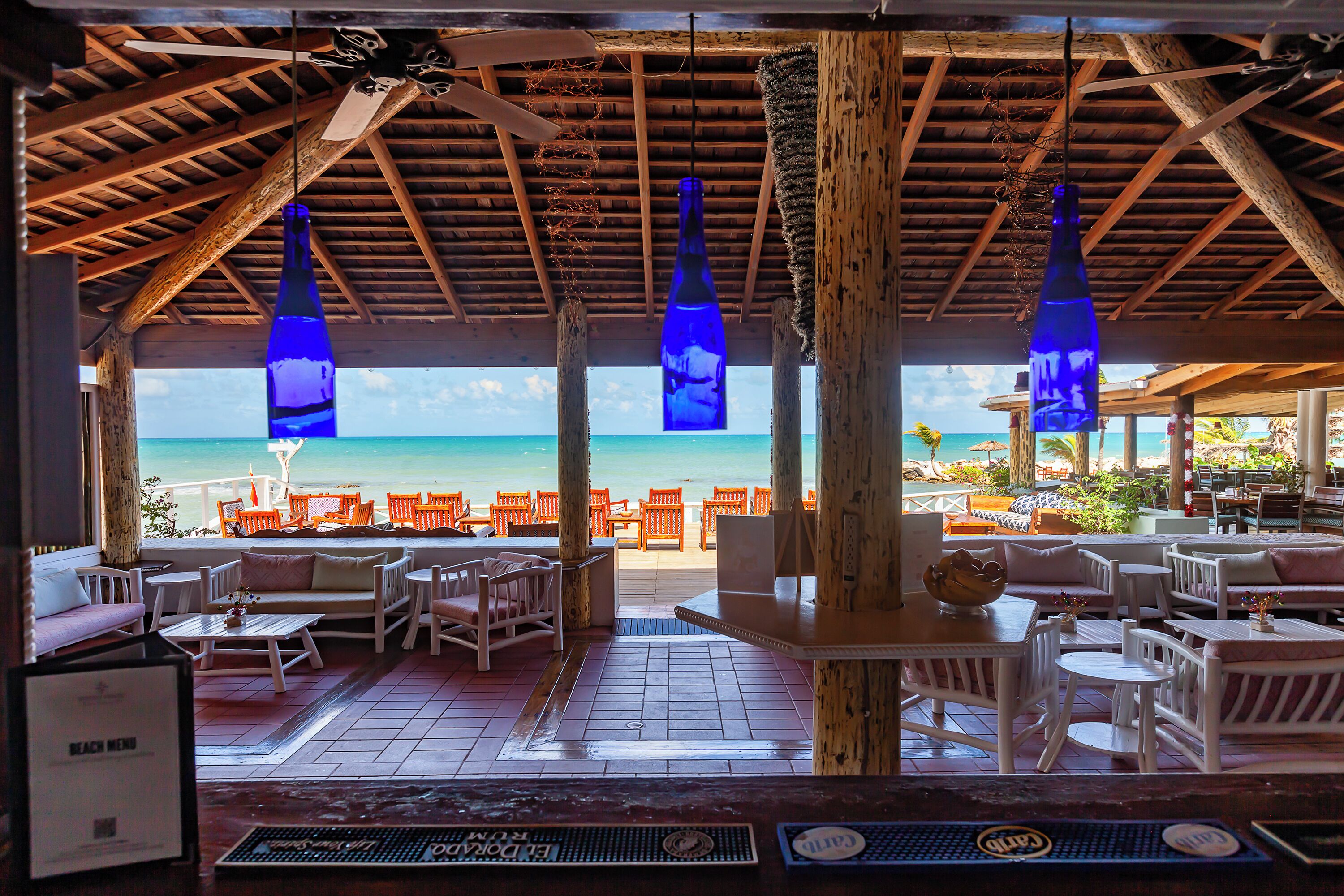 A beachside restaurant with wooden furniture and hanging blue bottles, overlooking the ocean.