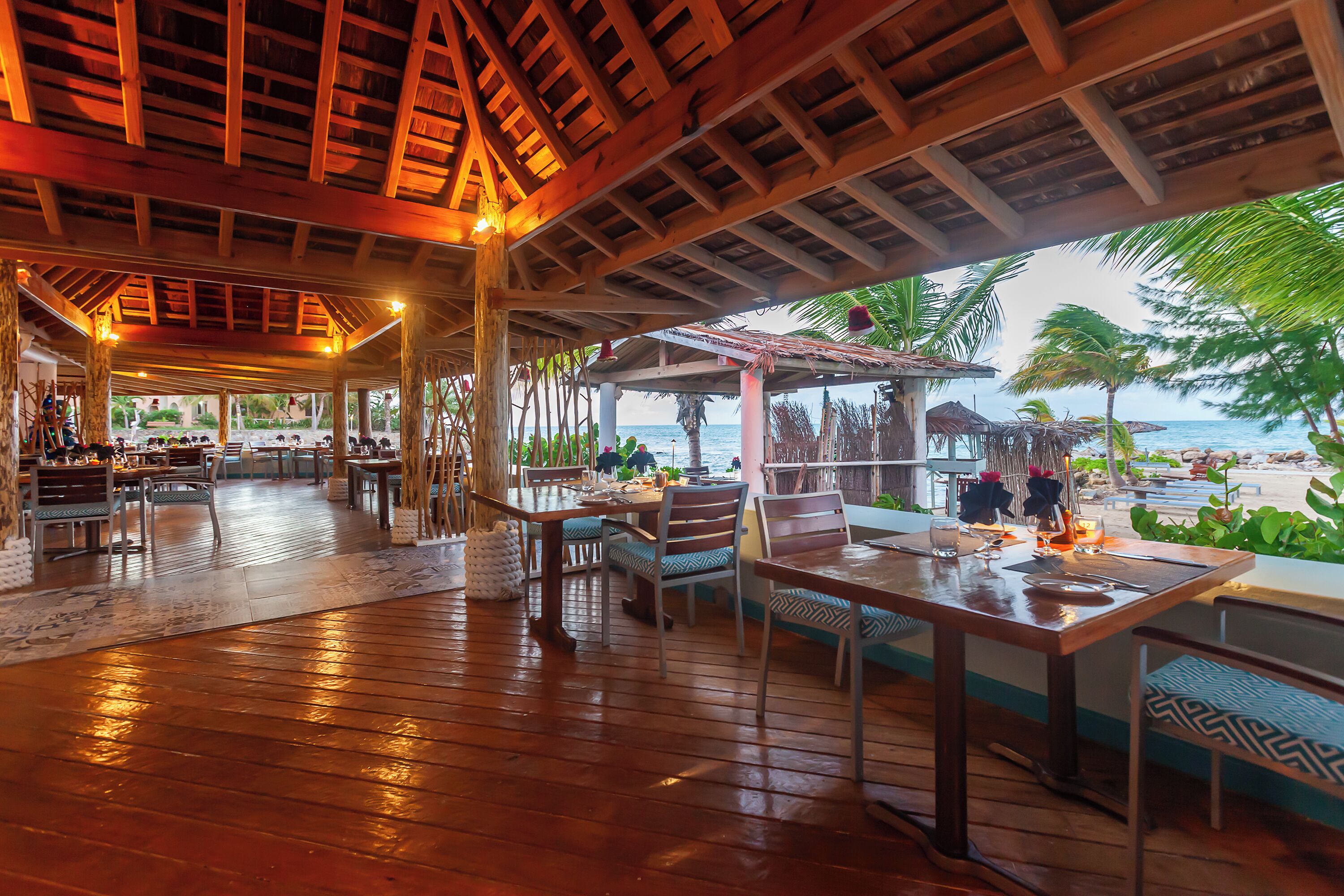 An open-air restaurant with wooden flooring and tables set for dining, overlooking a beach with palm trees and the ocean.