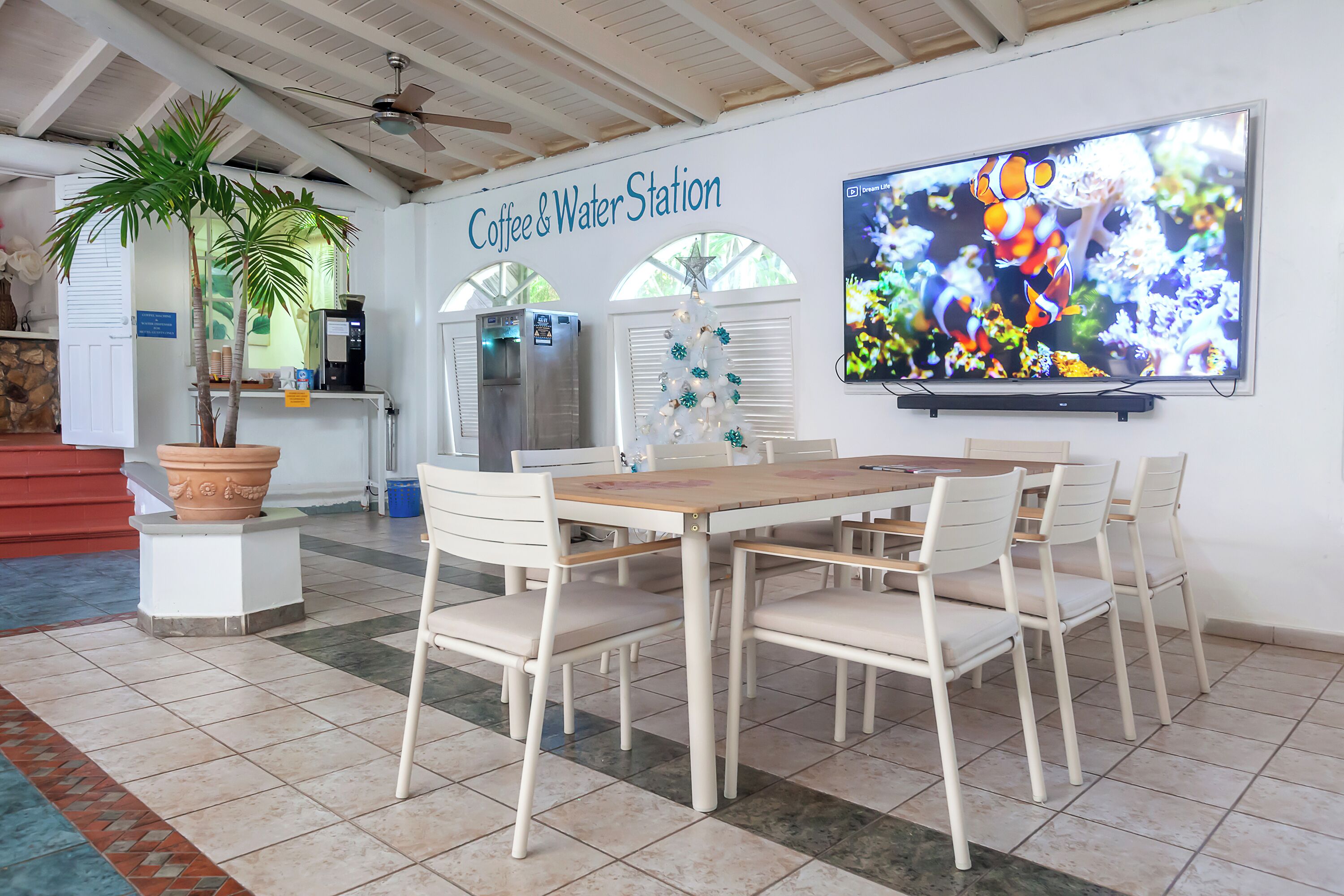 A Coffee & Water Station with a large TV, white chairs, a wooden table, and a potted plant.