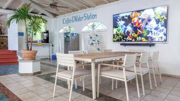 A Coffee & Water Station with a large TV, white chairs, a wooden table, and a potted plant.
