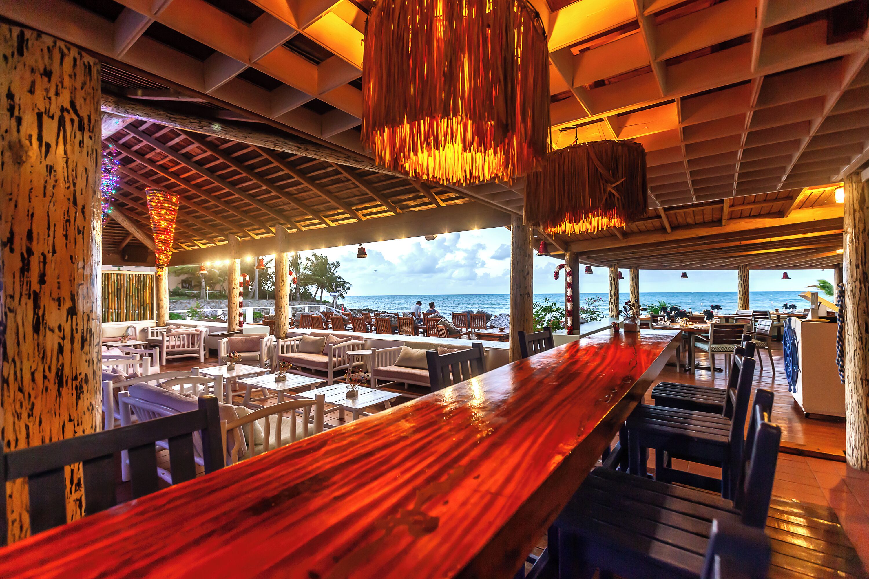 An outdoor restaurant with a wooden bar, seating area, and ocean view.