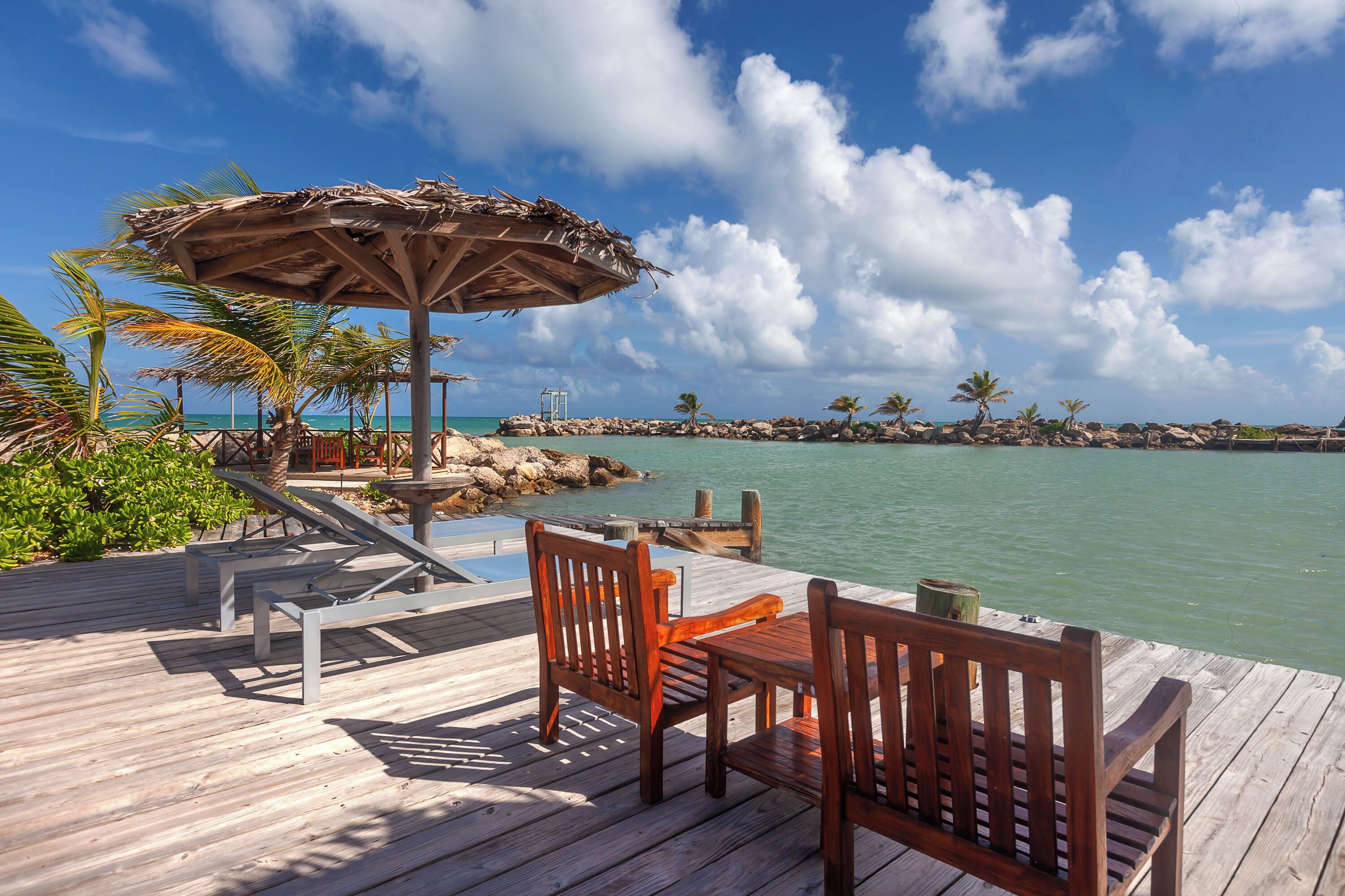 Eine Holzterrasse mit Tisch und Stühlen, einem Reetdachschirm und Blick auf das Meer und Palmen.