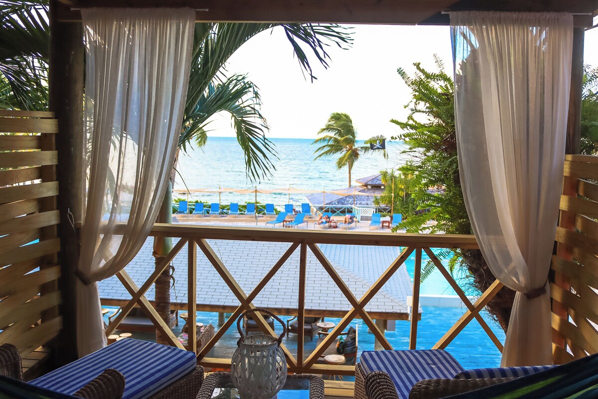 A balcony with wicker furniture, a view of the pool and beach, and a tropical setting.