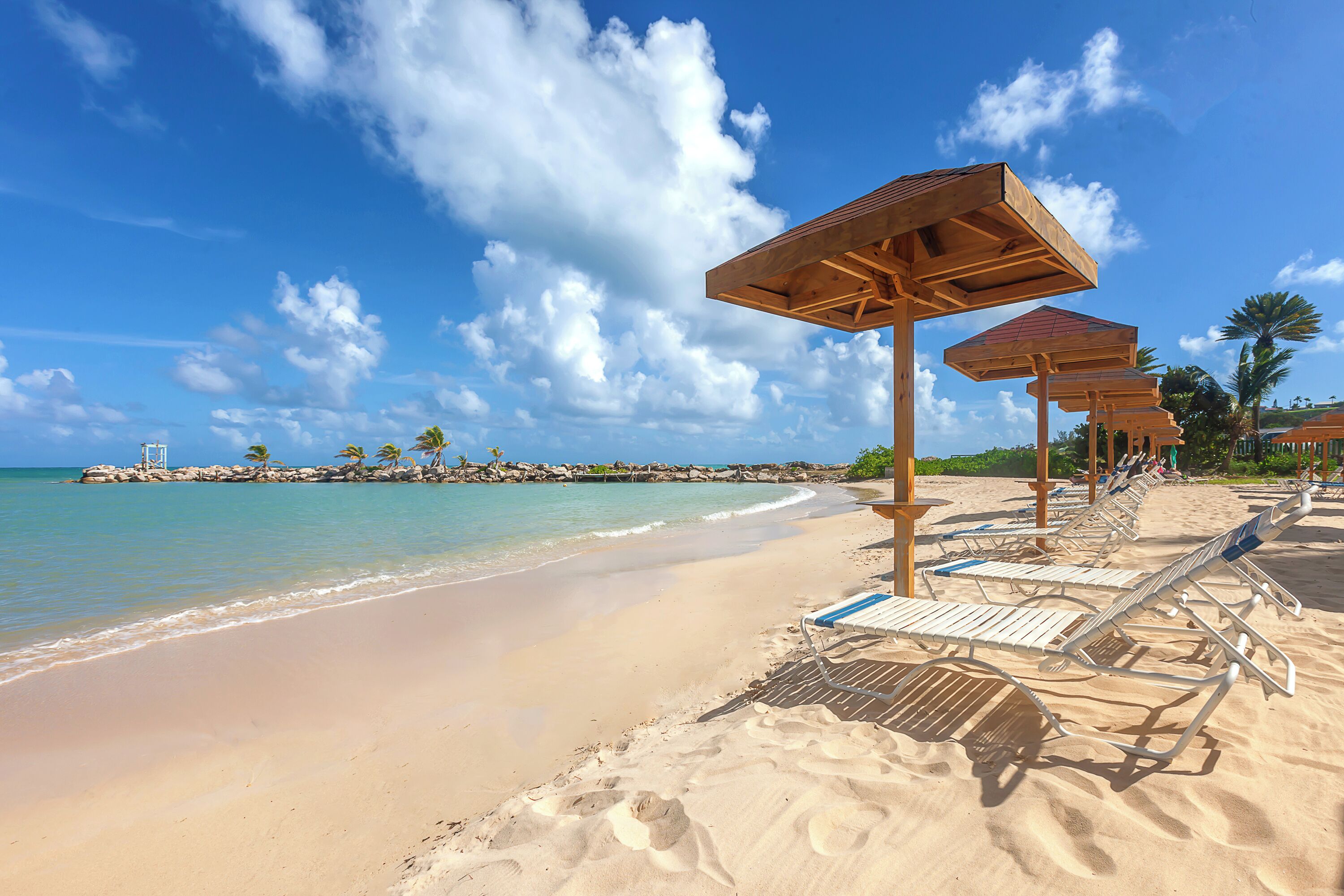 A beach with rows of sun loungers and wooden umbrellas, a clear blue sky, and a calm sea.