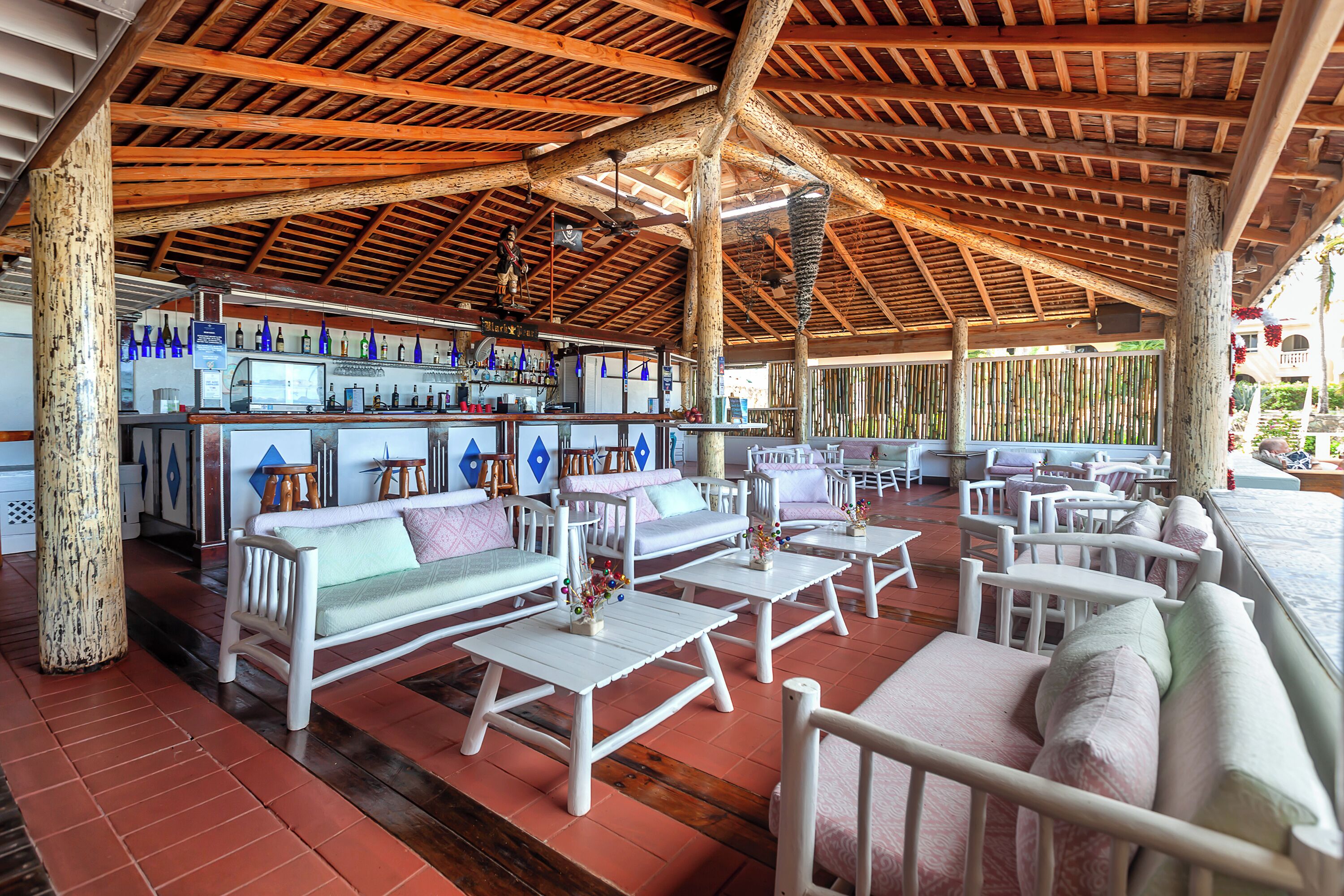 Uma área de estar coberta ao ar livre com mesas de madeira e sofás brancos, um bar com garrafas azuis e uma cerca de bambu ao fundo.