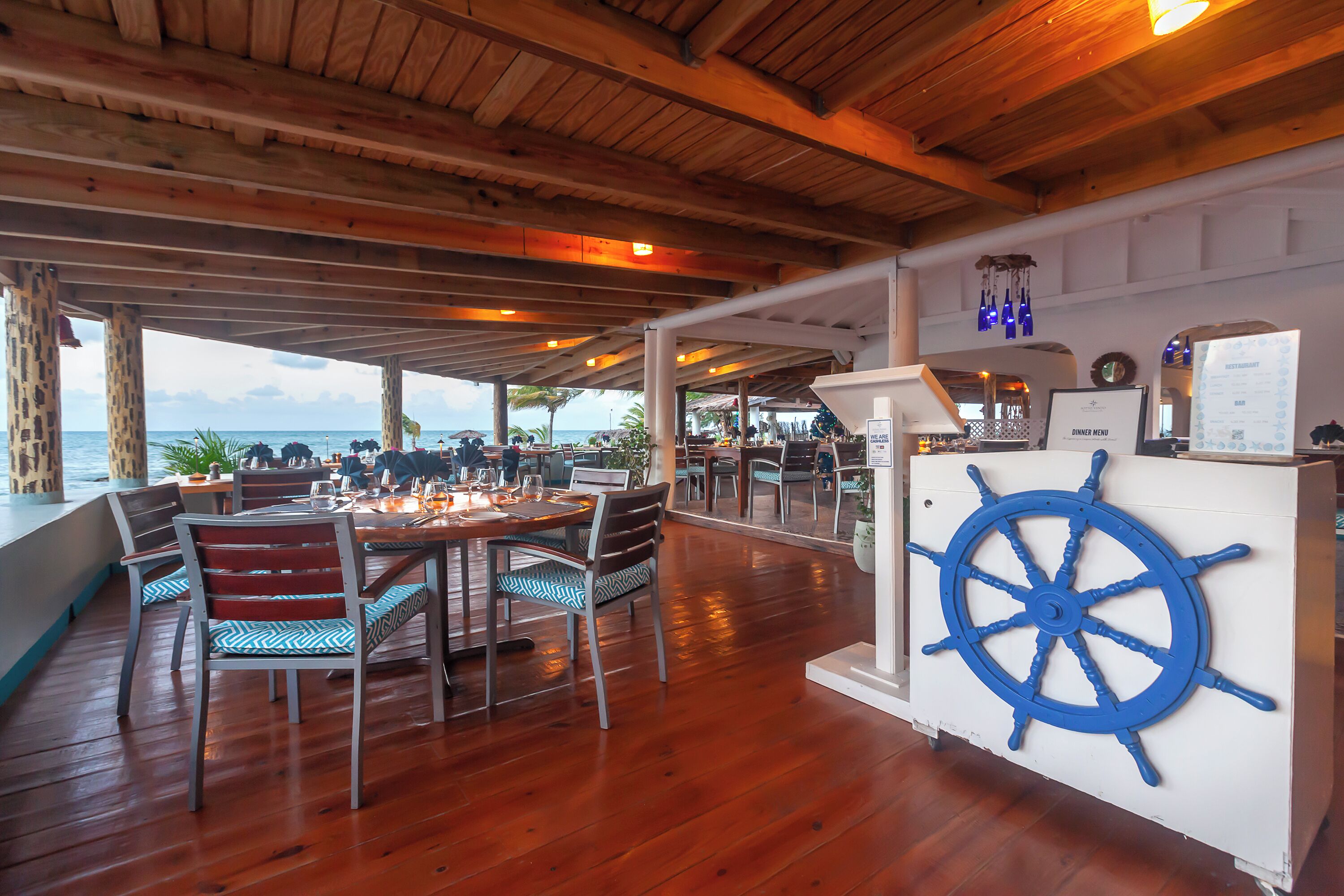 An outdoor dining area with wooden tables and chairs, a ship's wheel decoration, and a menu board.
