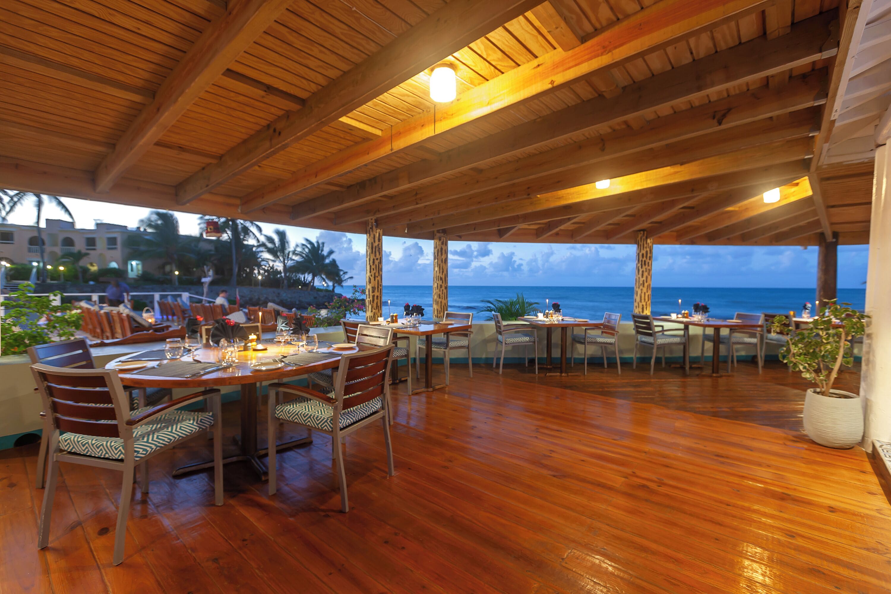 An outdoor dining area with wooden tables and chairs, overlooking the ocean.