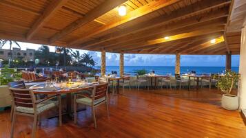An outdoor dining area with wooden tables and chairs, overlooking the ocean.