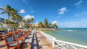 A wooden deck with outdoor seating, overlooking a beach with palm trees and a clear blue sky. - Ocean Point Beach Resort & Spa - Adults Only (St. John's)