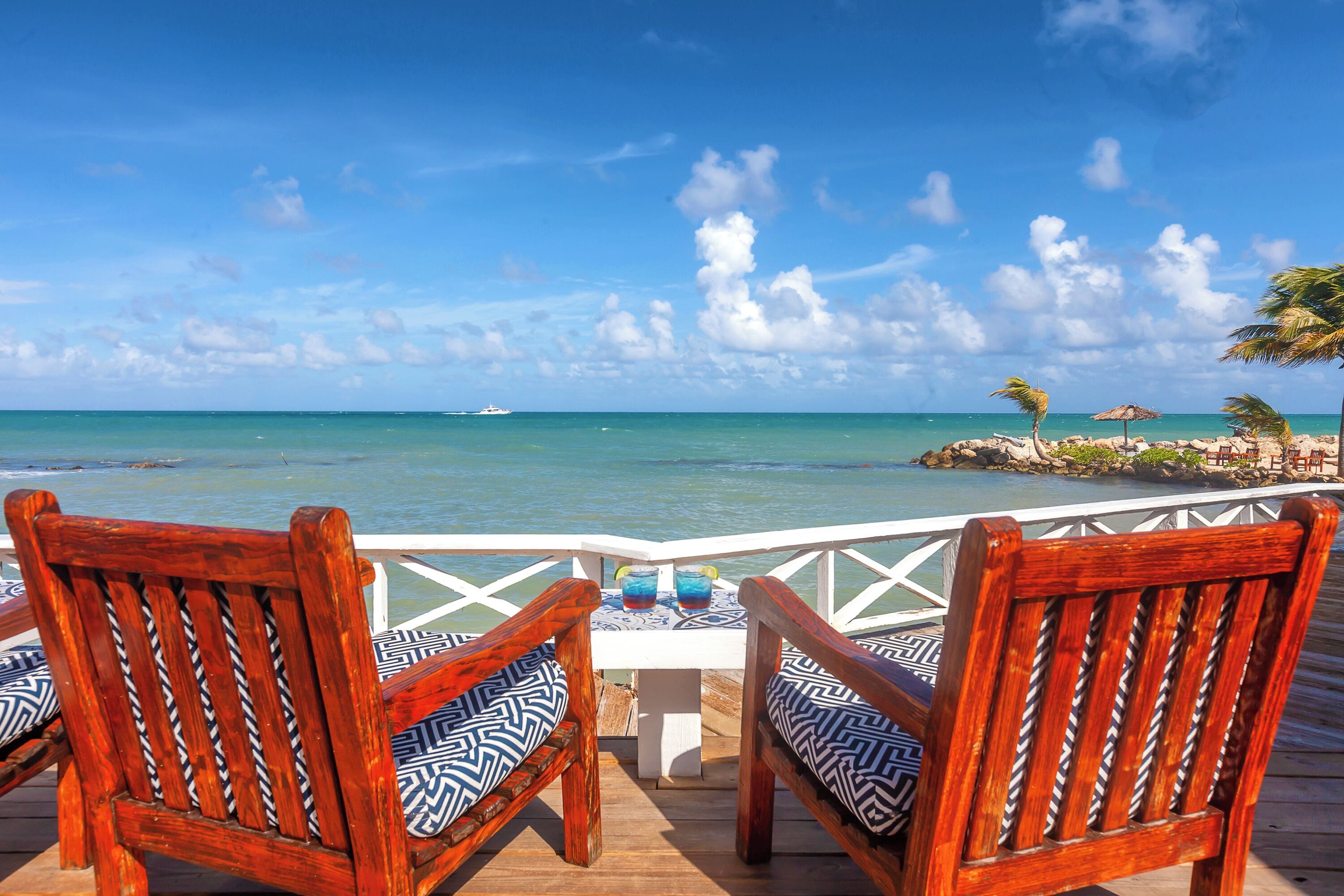 A wooden deck with two chairs and a table, overlooking a beach with a clear view of the ocean and a boat in the distance.