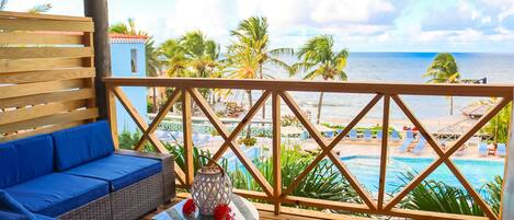 A wooden deck with a blue sofa and a small table with a decorative item. In the background, there is a pool and a beach with palm trees and the ocean.
