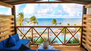 A wooden deck with a blue sofa and a table, overlooking a beach with palm trees and a pool. - Ocean Point Beach Resort & Spa - Adults Only (St. John's)