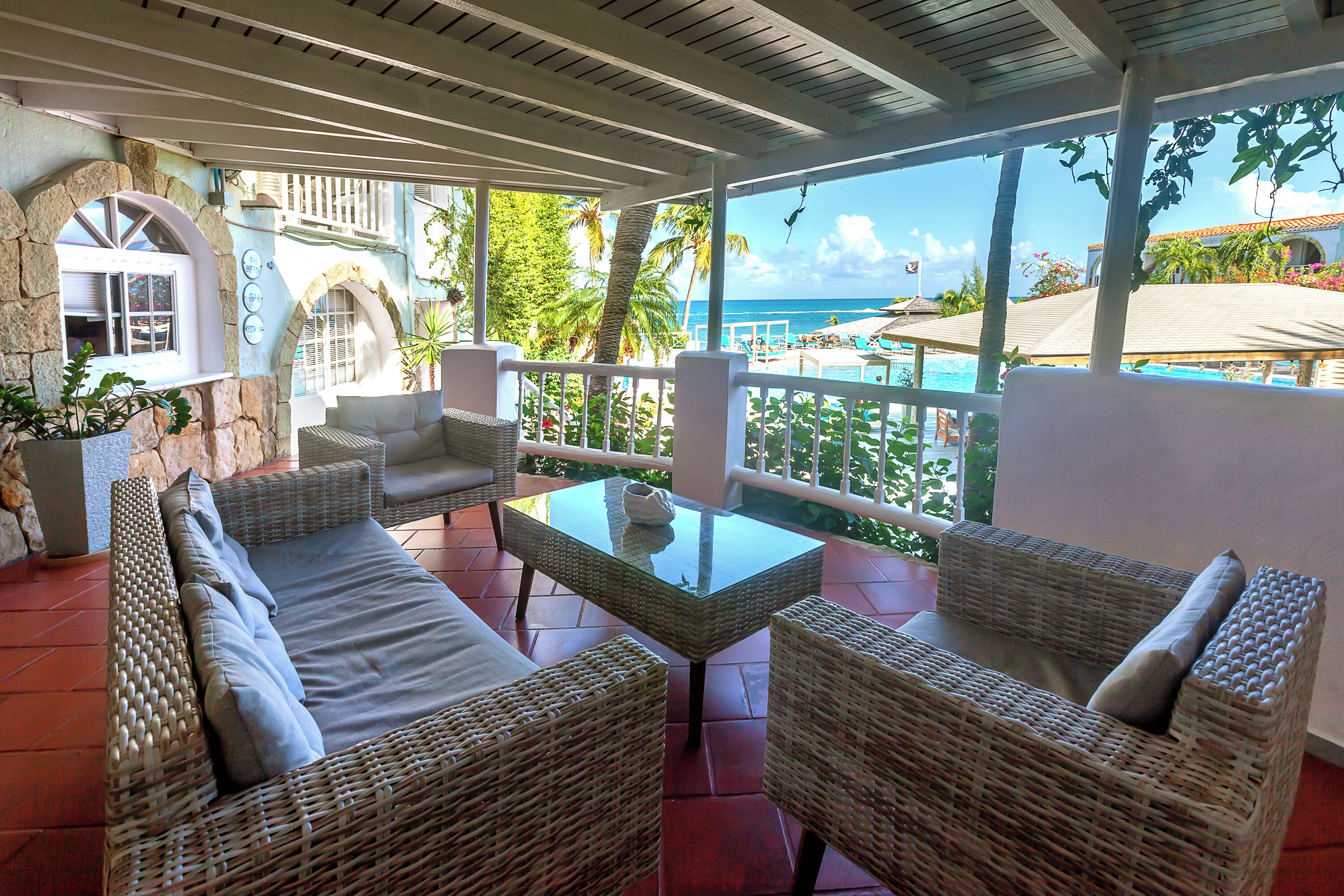 Un espacio exterior cubierto con mobiliario de mimbre y una mesa de cristal, con vistas a una piscina y palmeras.