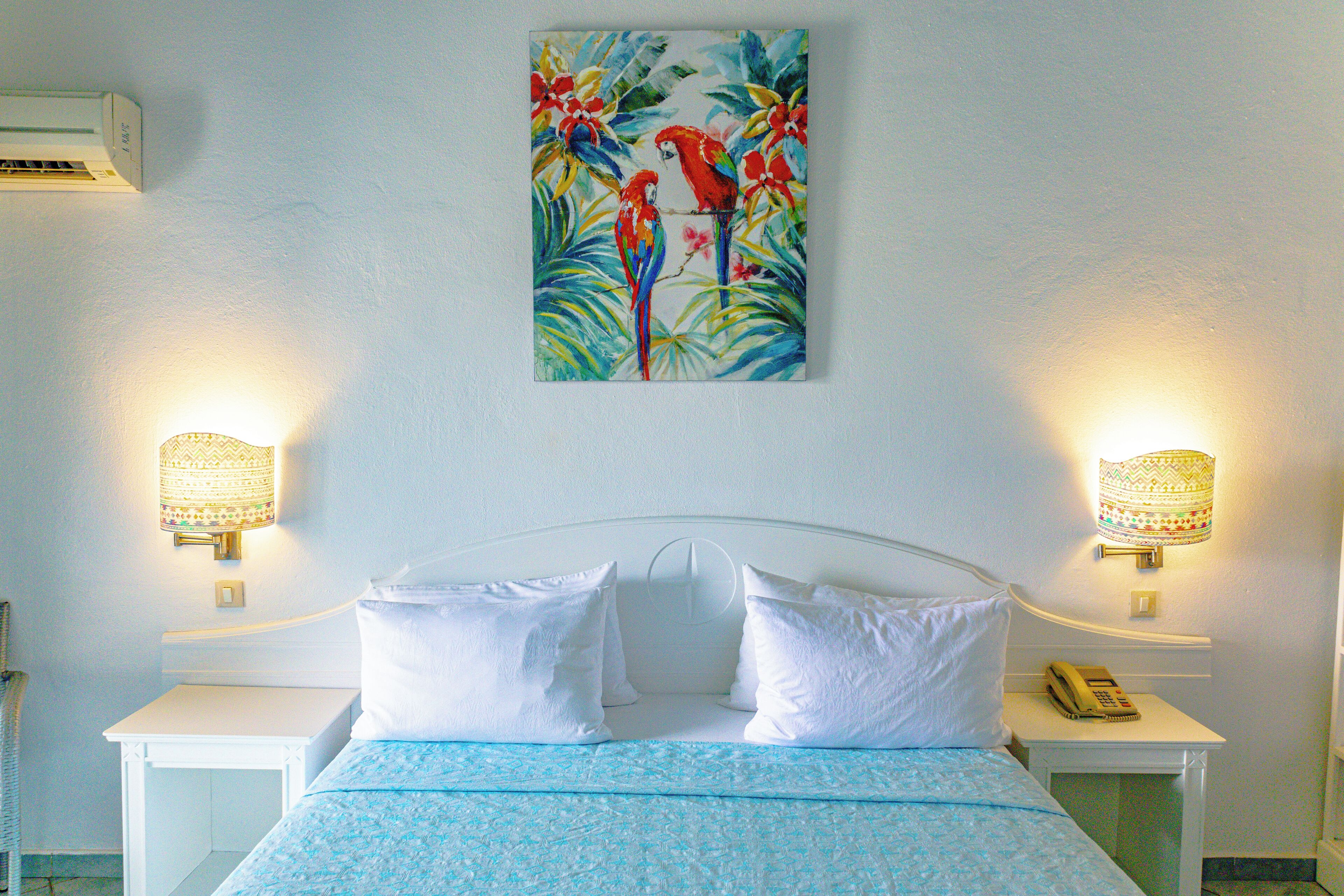 A hotel room with a bed, white nightstands, a telephone, and a wall-mounted painting of parrots.