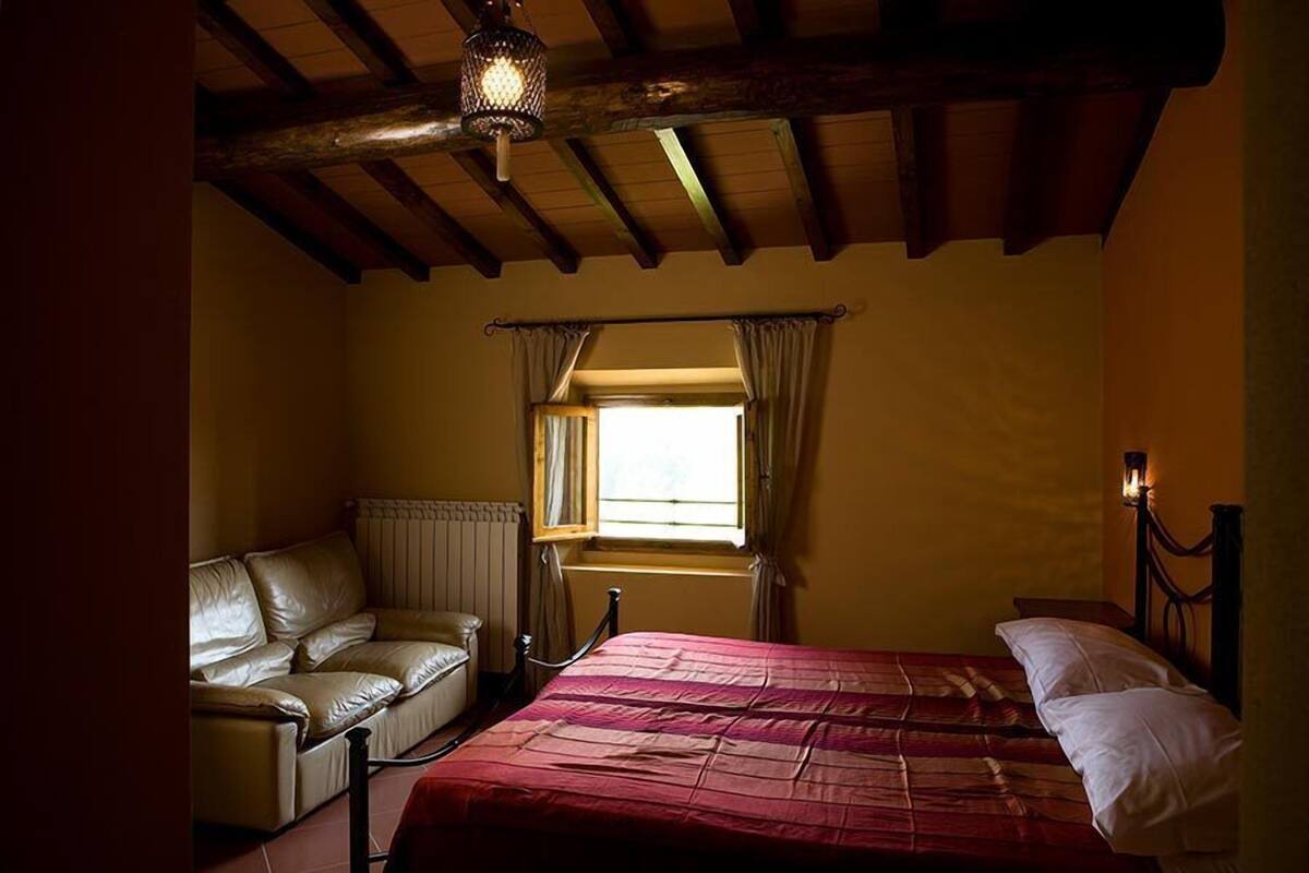 A bedroom with a bed, a leather armchair, a window with curtains, and a wooden ceiling beam.