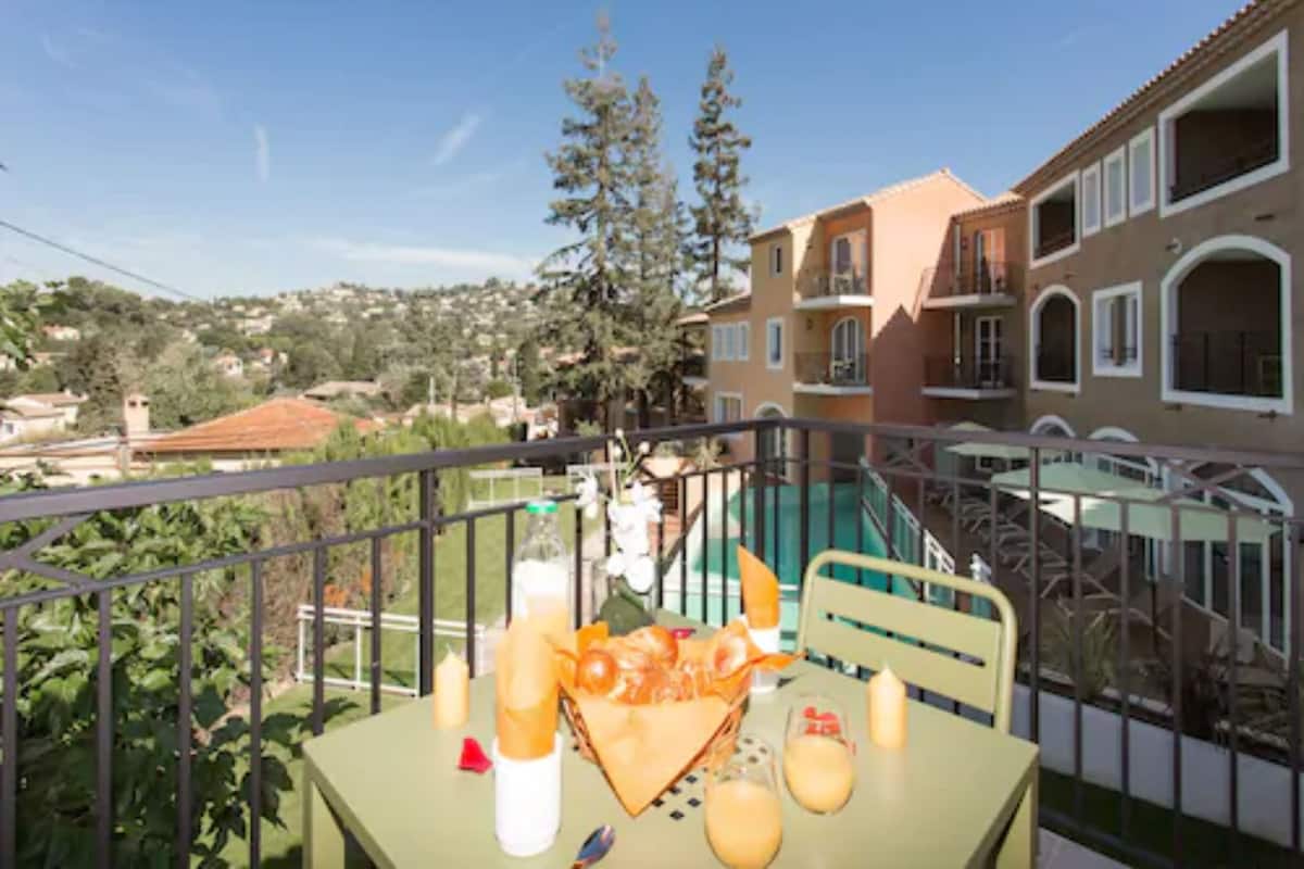 Un balcón con una mesa lista para el desayuno, con vista a una zona residencial con casas y árboles.