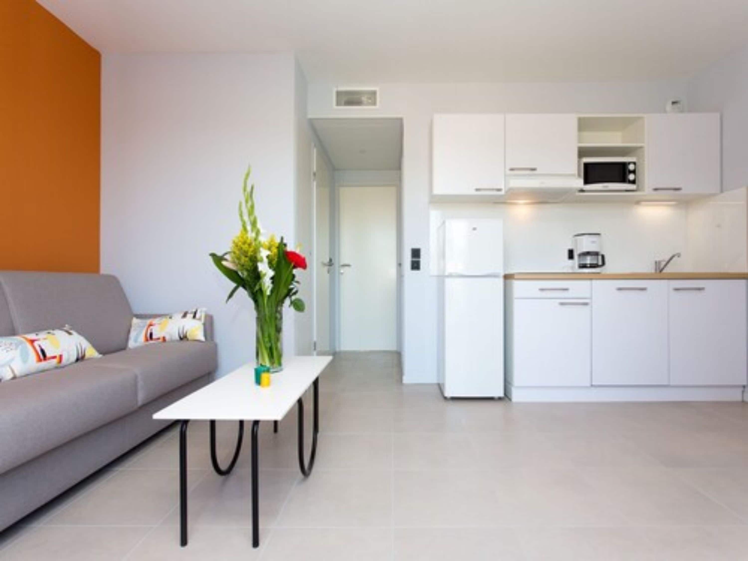 Un salon moderne avec un canapé gris, une table basse blanche et un vase rempli de fleurs. On aperçoit également une cuisine avec des meubles blancs, un four à micro-ondes et une machine à café.