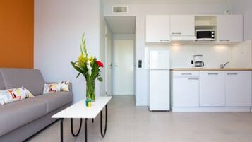 Un salon moderne avec un canapé gris, une table basse blanche et un vase rempli de fleurs. On aperçoit également une cuisine avec des meubles blancs, un four à micro-ondes et une machine à café.