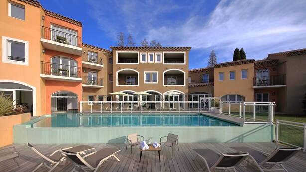 A poolside area with lounge chairs and a table, surrounded by multi-story buildings with balconies and a clear blue sky. - Adonis Mougins (Mougins)