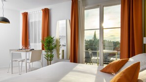 A hotel room with a large bed, a desk with a chair, a potted plant, and a view of a cityscape through a large window. - Adonis Mougins (Mougins)