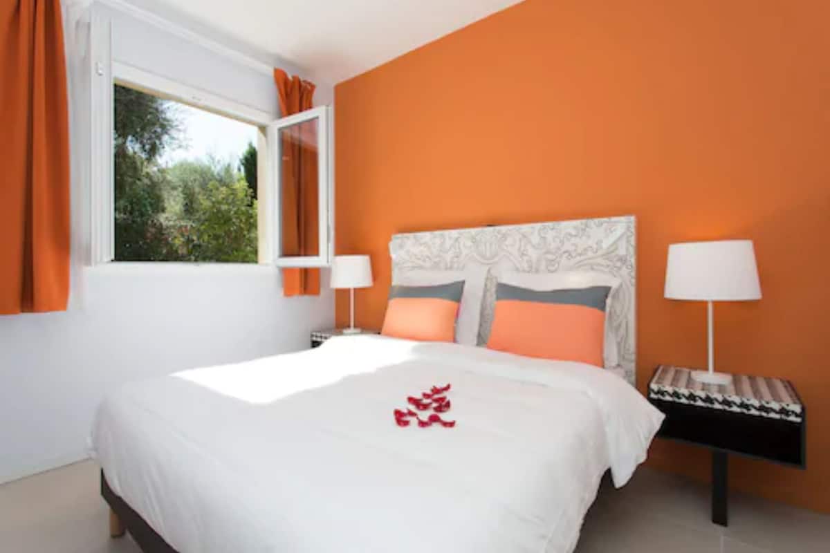 Un dormitorio con una cama grande, dos mesitas de noche con lámparas, una pared naranja y una ventana con cortinas.