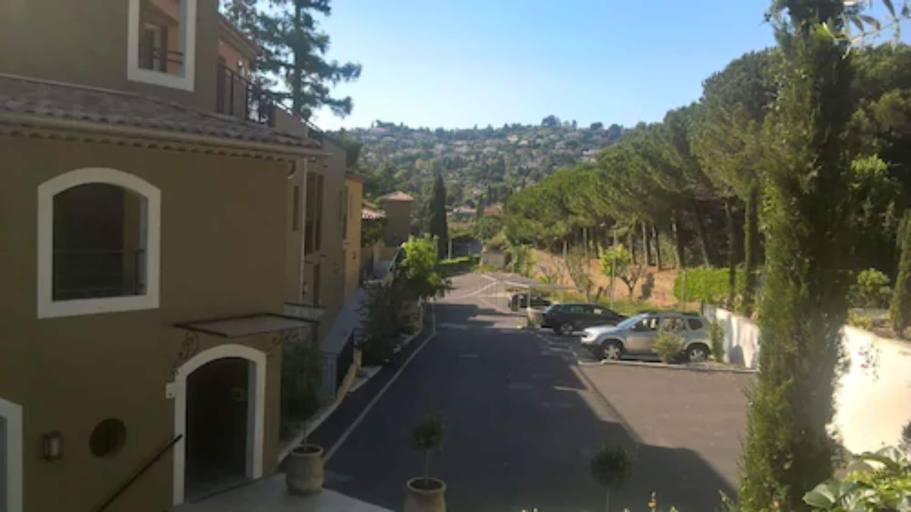 A residential street with houses, parked cars, and a hilly landscape in the background.