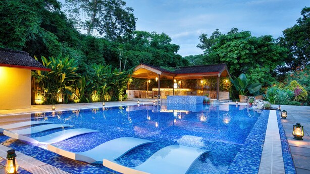 A swimming pool with a tiled deck, surrounded by lush greenery and a wooden structure with seating. - GREEN MANSIONS JUNGLE RESORT (Sauraha)