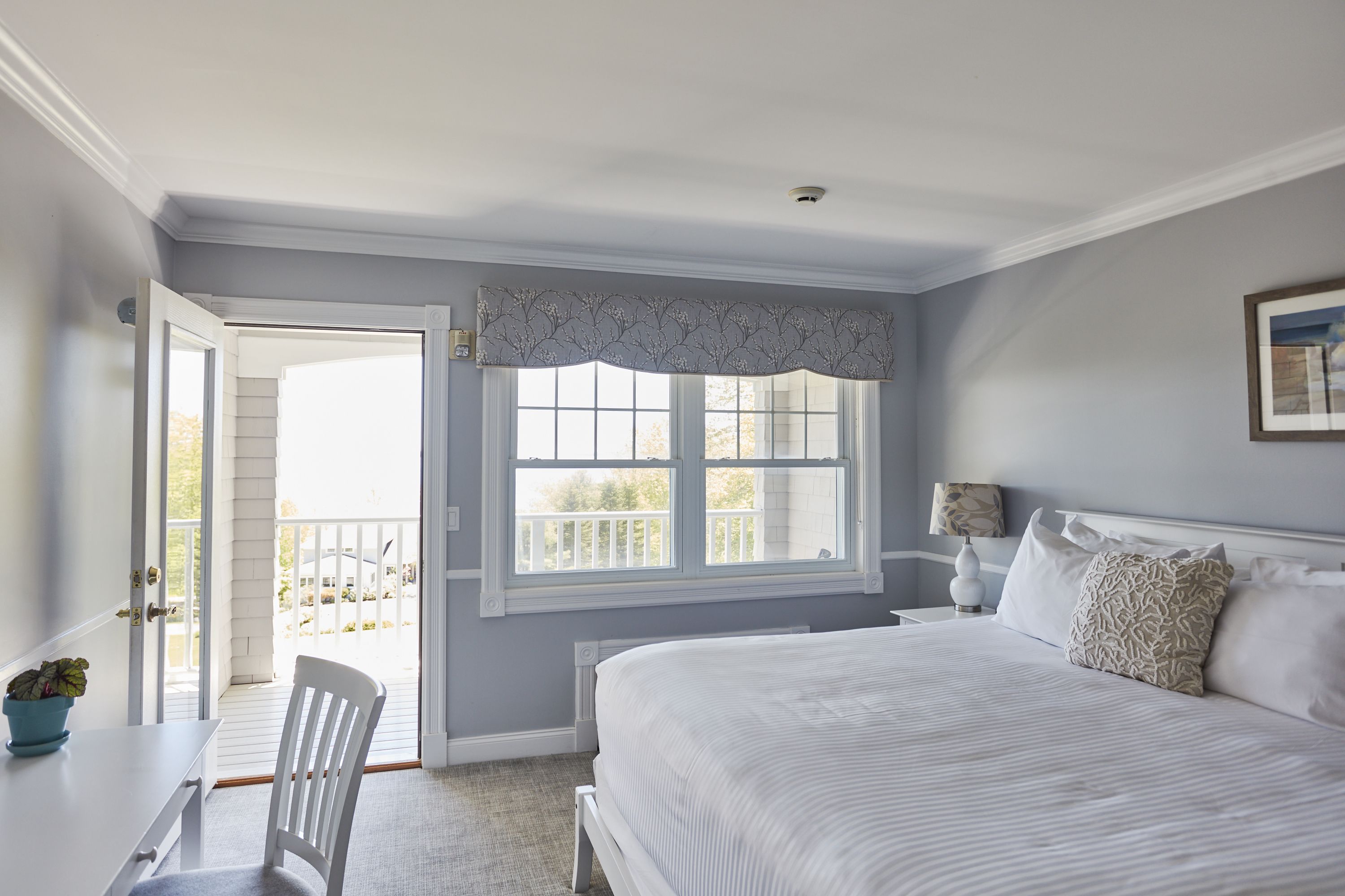 A bedroom with a large bed, a window with a view, a door leading to a balcony, and a small table with a plant.