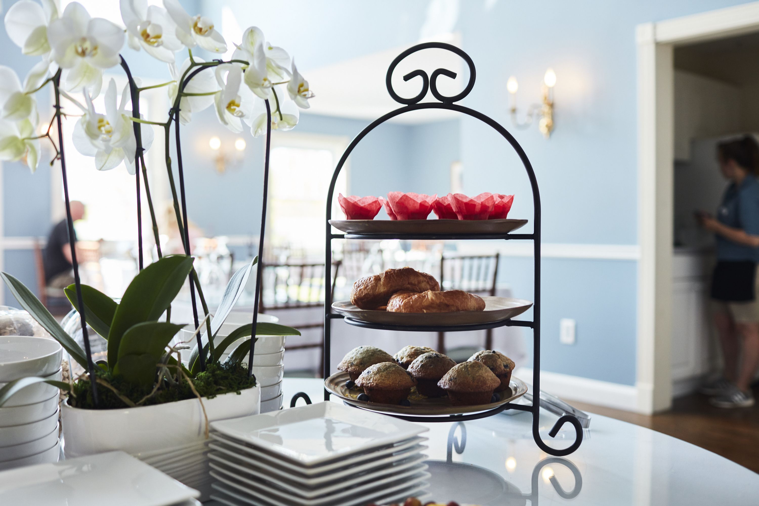 Un présentoir à trois niveaux, orné de pâtisseries et surmonté d’une plante en pot, est posé sur une table.