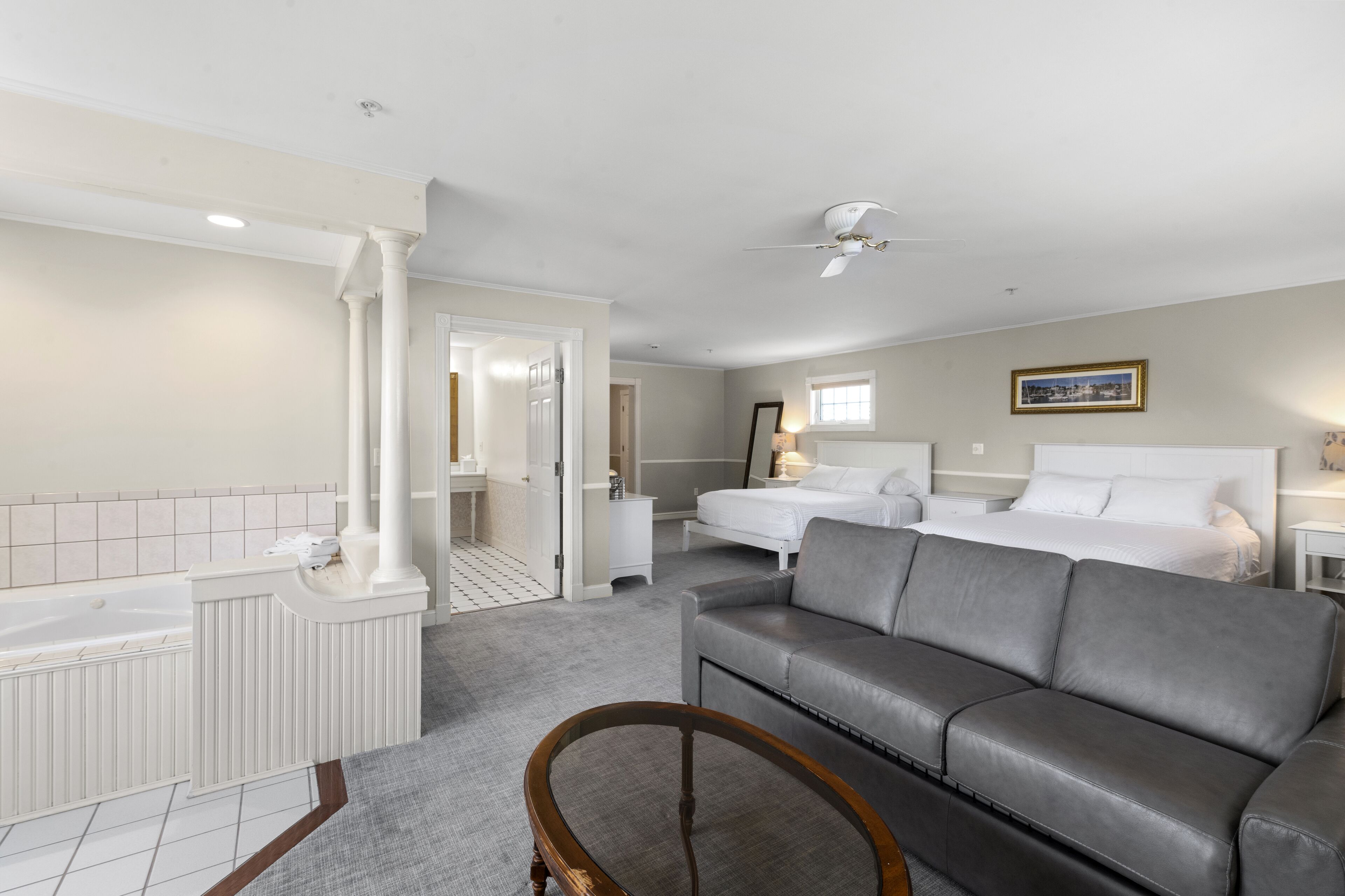 A hotel room with a bathtub, two beds, a gray sofa, a wooden chair, and a ceiling fan.