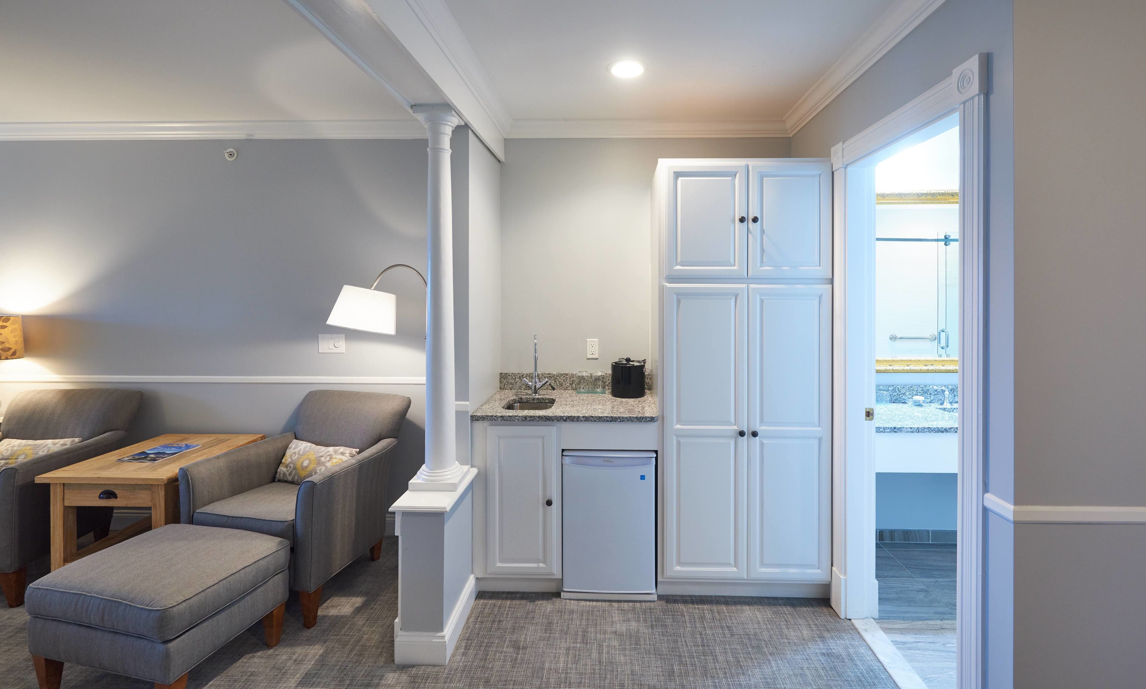 A compact living space with a kitchenette, grey upholstered seating, a small refrigerator, and a view through an open door.