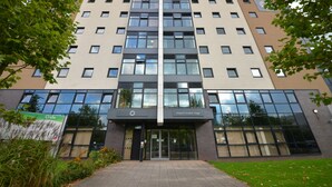 Un edificio de varios pisos con un diseño arquitectónico moderno, que presenta una entrada central con puertas de vidrio y un cartel que indica que es una residencia universitaria.