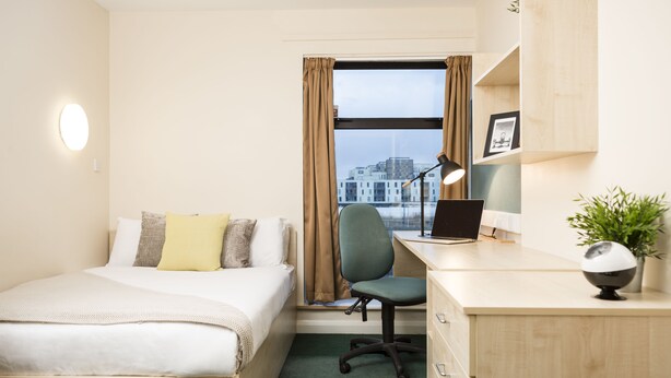 Habitación de hotel con cama, escritorio, silla, ordenador portátil y vistas a edificios a través de una ventana.