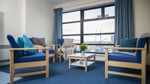 Una habitación con dos sillas tapizadas en azul, una mesita de madera y vistas a un paisaje urbano a través de amplias ventanas.