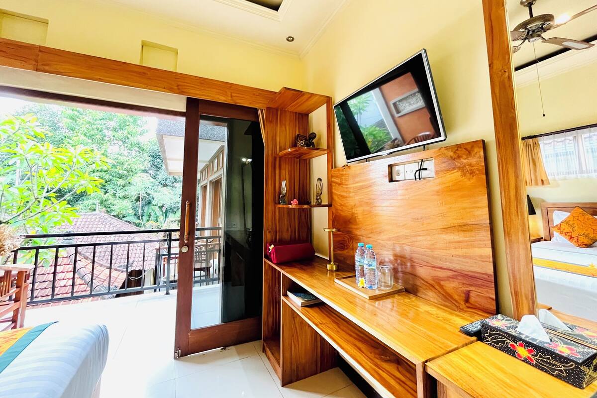 A room with a wooden desk, a TV mounted on the wall, a bed with a view of the outdoors, and a balcony with a railing.