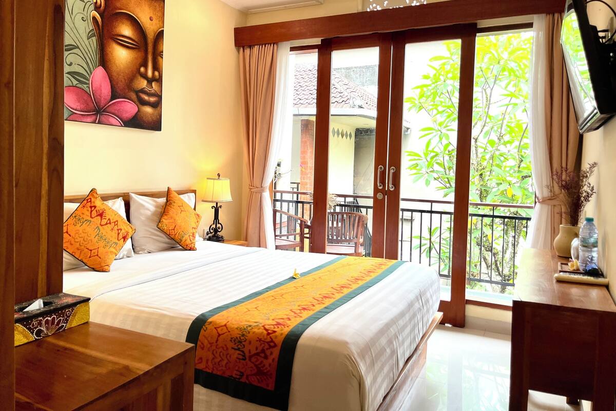A hotel room with a bed, a nightstand, a TV, a balcony with a view of greenery, and a painting on the wall.