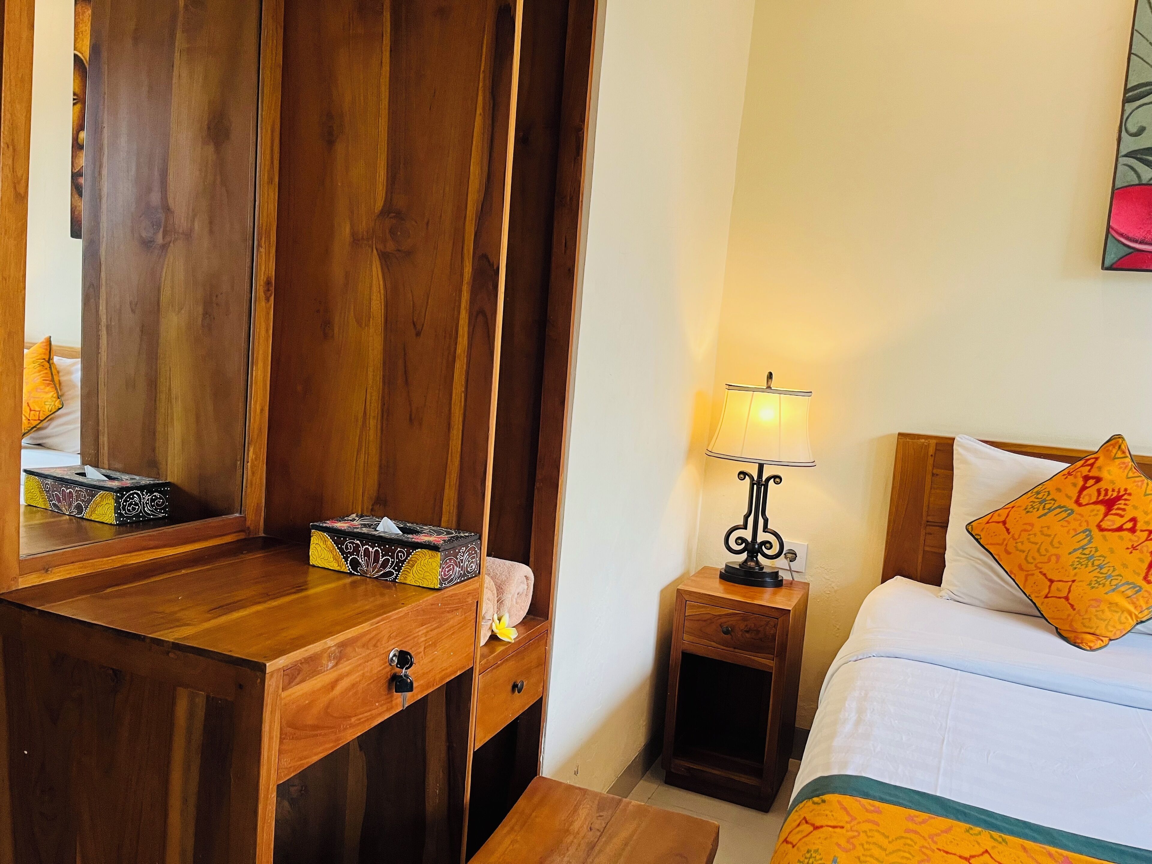 A wooden dresser with a mirror, a bedside table with a lamp, a bed with a patterned pillow, and a small nightstand.