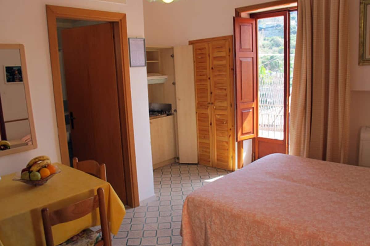 A bedroom with a bed, a table with a fruit bowl, a wardrobe, and a window with curtains.