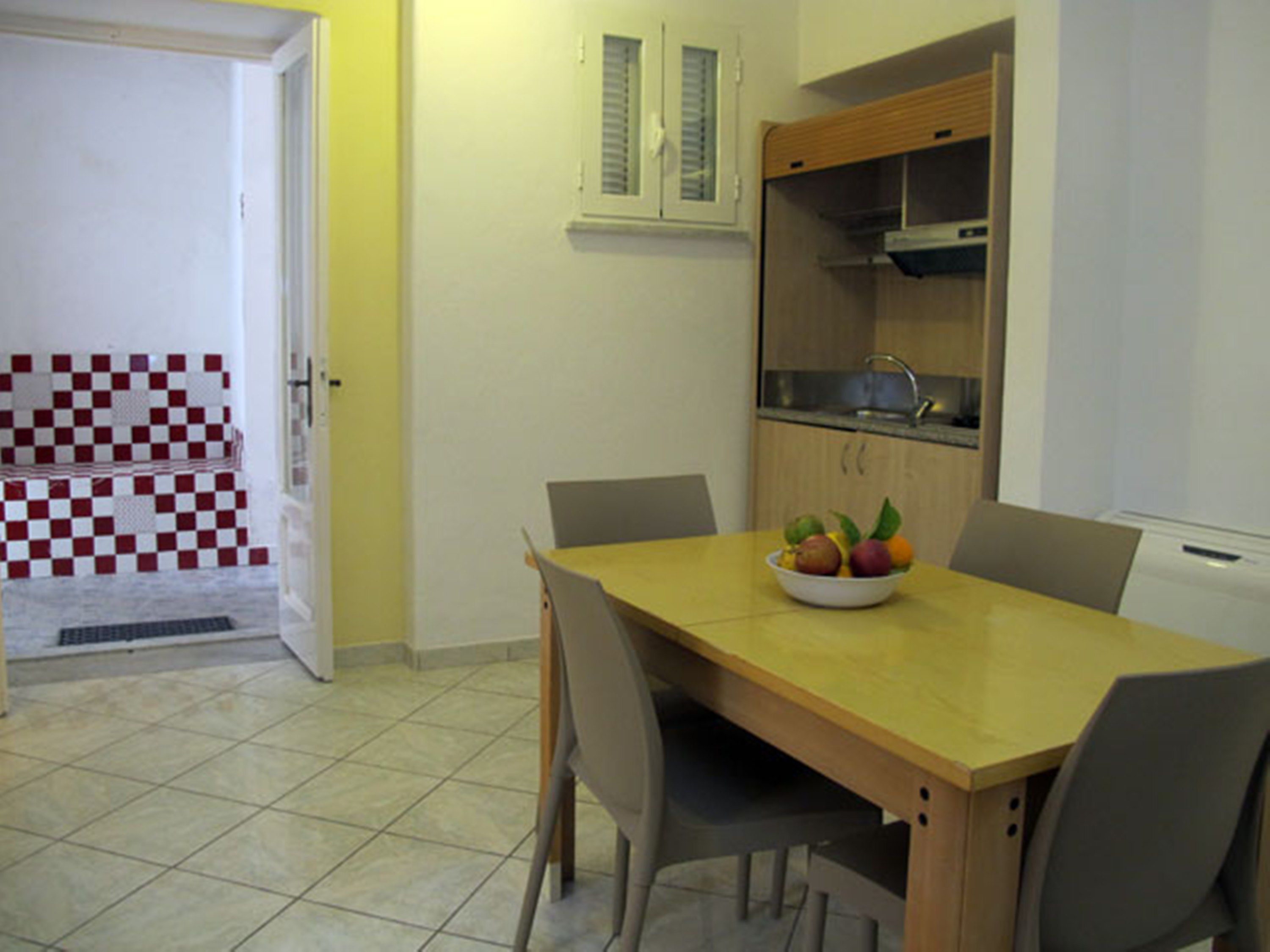 A dining area with a wooden table, four chairs, and a bowl of fruit. There is a kitchen area with a sink and cabinets.