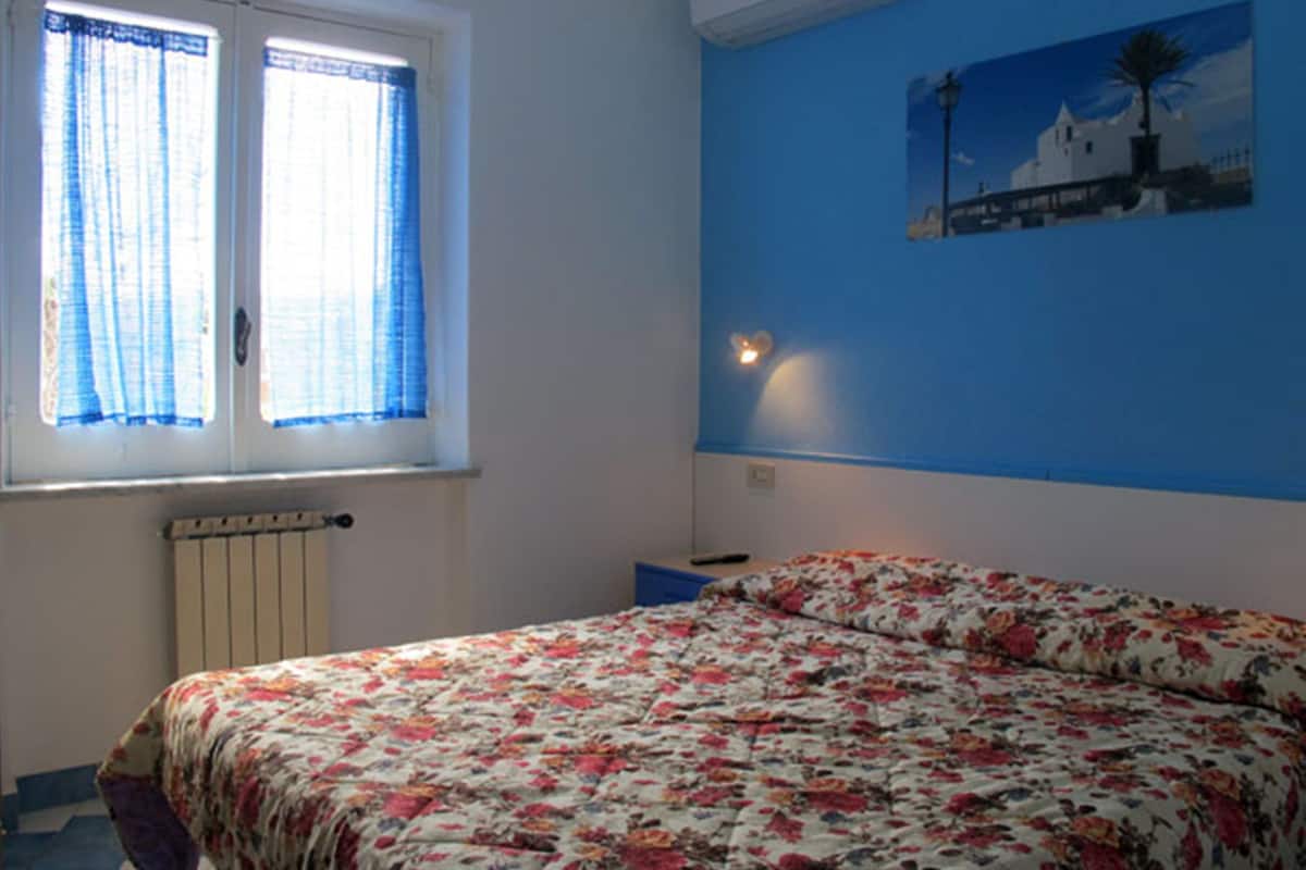 A bedroom with a floral bedspread, a window with blue curtains, a wall-mounted air conditioner, and a painting of a white building.