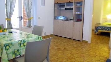 A dining area with a table covered by a green tablecloth, chairs, a kitchen area with cabinets, and a bedroom with a bed and a nightstand.