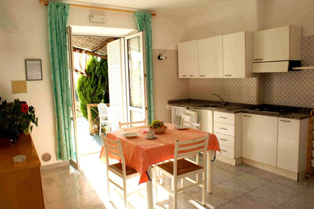 A compact kitchen with white cabinets, a dining table with a tablecloth, and a view through an open door to a garden.