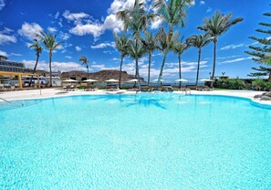 Una piscina azul cristalina rodeada de palmeras y sillas de descanso, con una montaña al fondo.