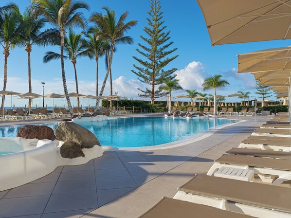 A pool area with lounge chairs, umbrellas, and palm trees. - H10 Costa Mogán (Mogan)