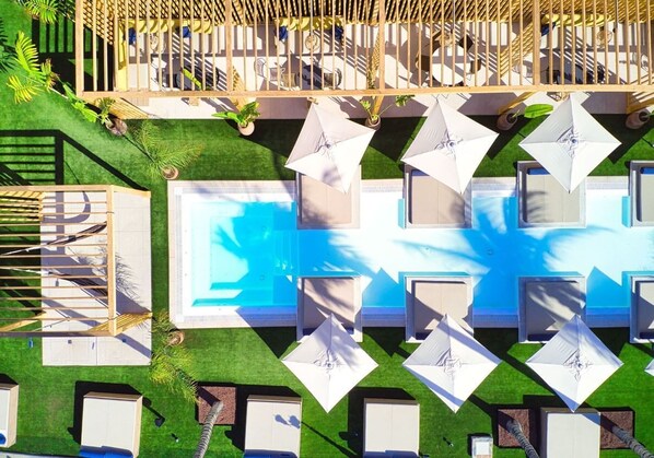 Vista aérea de un hotel con piscina rodeada de tumbonas y sombrillas, una terraza de madera y un área cercada con asientos.