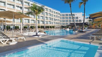 Zona de piscina de hotel con múltiples sillas de descanso y sombrillas, una piscina grande y un edificio con varios balcones.