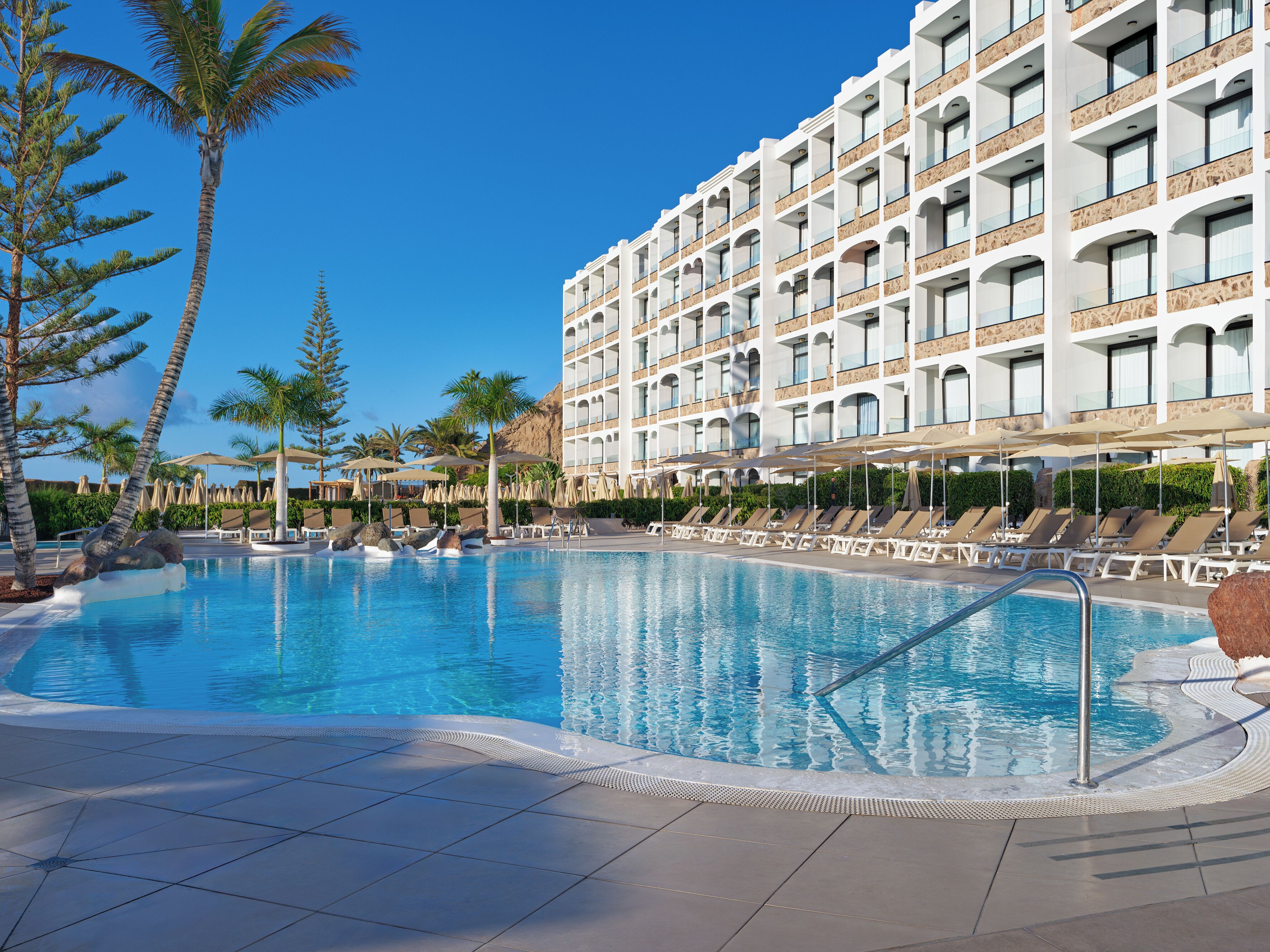 Un hotel con una piscina grande, tumbonas y palmeras.