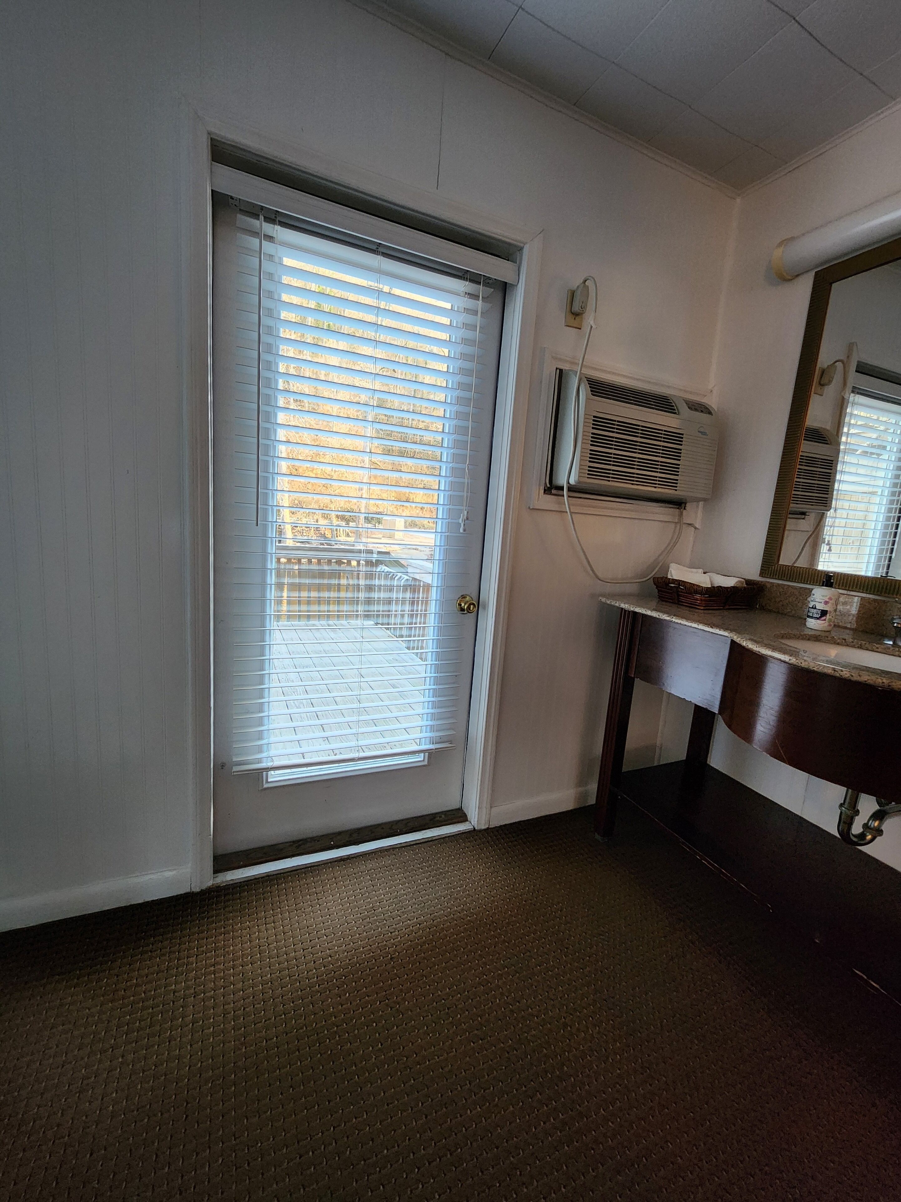 A room with a closed door, a window with blinds, a wall-mounted air conditioner, a sink, and a mirror.