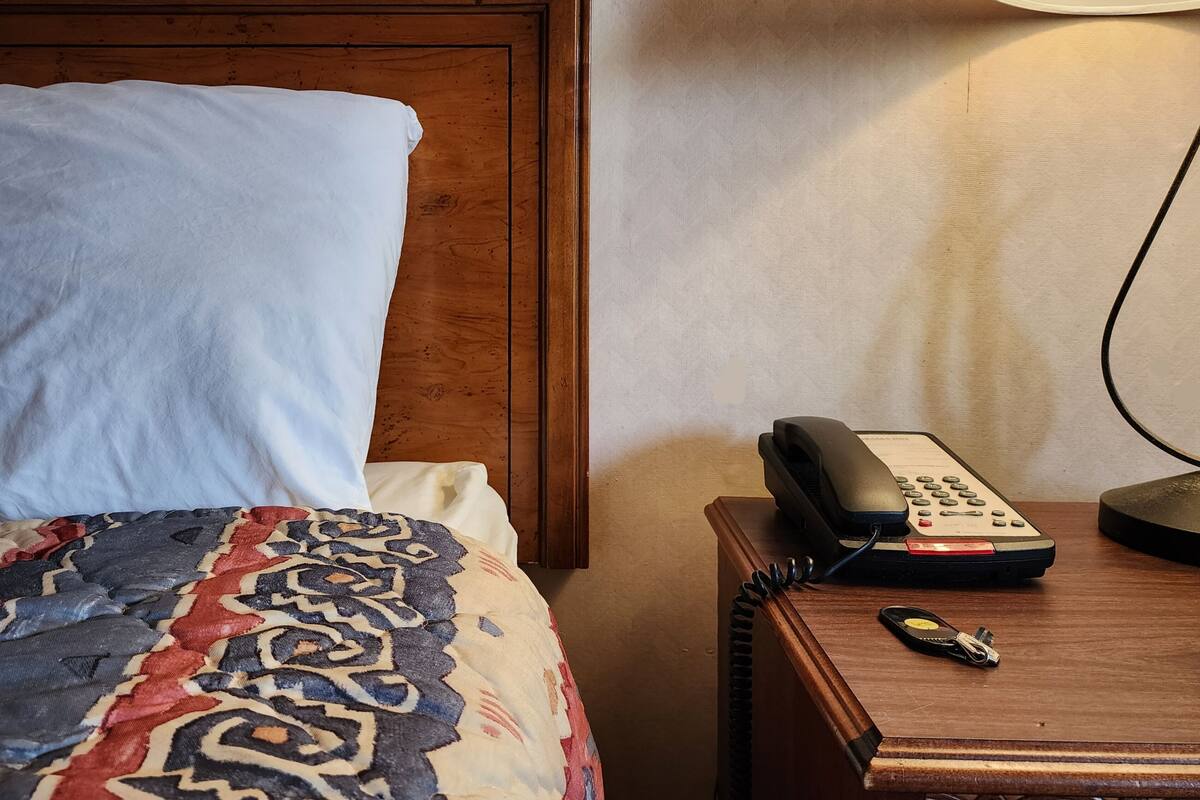 A hotel room with a wooden headboard, a bed with patterned bedding, a bedside table with a telephone and keys, and a lamp on the wall.