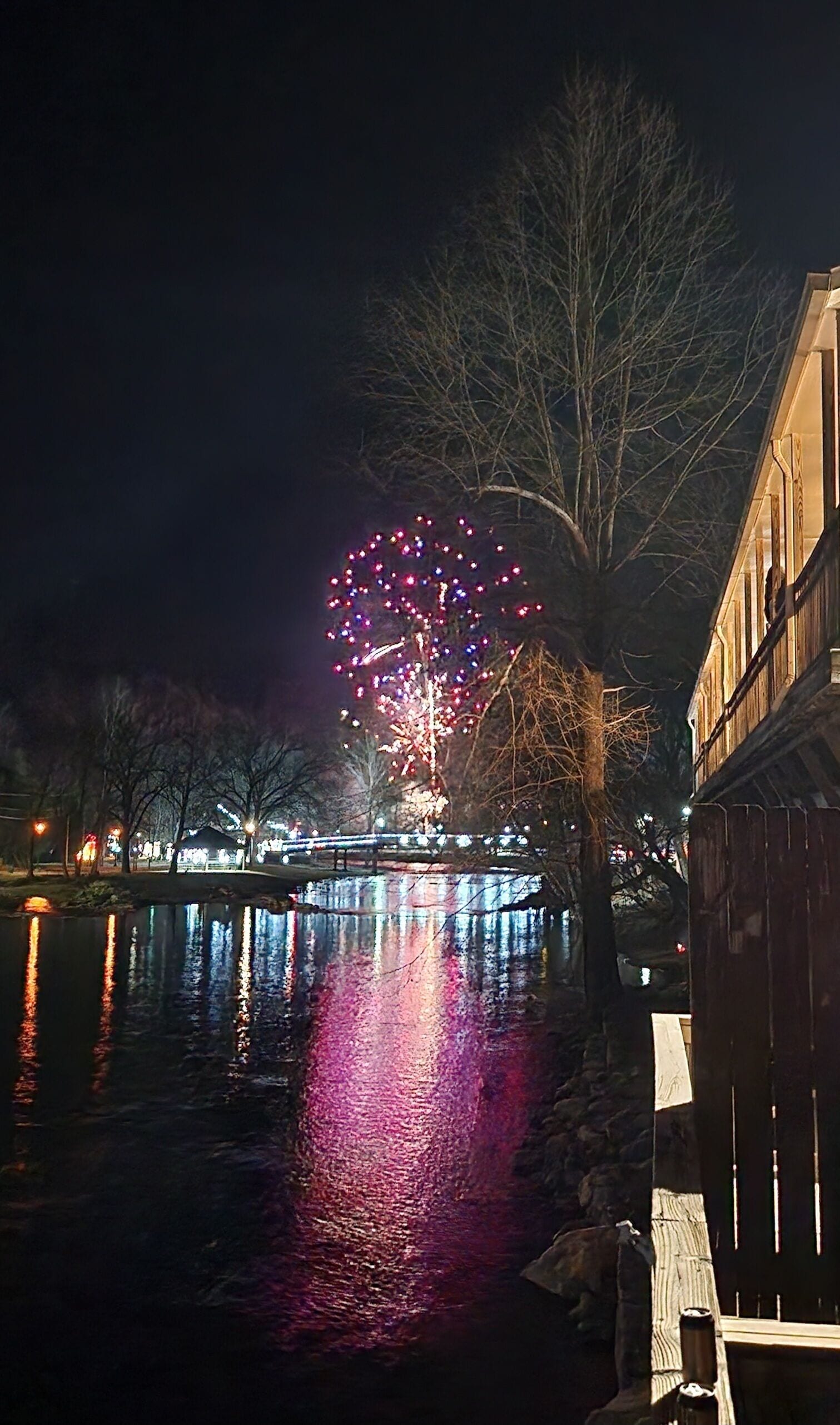 A nighttime view of a river with fireworks reflection, a bridge, and buildings along the riverbank.